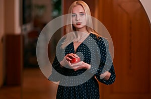 Young attractive blonde woman with short hair posing in a black dress with white polka dots indoors, holding a red apple