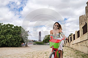 Young, attractive, blonde woman, wearing floral shirt, white pants and sunglasses, riding an electric bicycle for rent. Concept