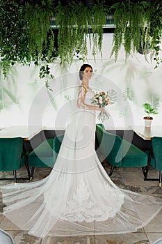 Young attractive bride in wedding dress with bridal veil looks at bouquet and smiles. Girl in wedding gown in modern