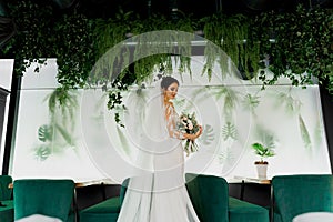 Young attractive bride in wedding dress with bridal veil looks at bouquet and smiles. Girl in wedding gown in modern