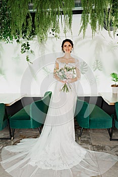 Young attractive bride in wedding dress with bridal veil looks at bouquet and smiles. Girl in wedding gown in modern