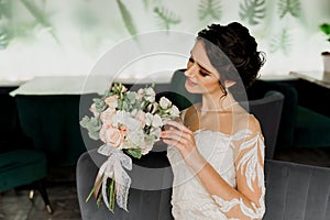 Young attractive bride in wedding dress with bridal veil looks at bouquet and smiles. Girl in wedding gown in modern