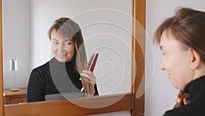 Young attractive brown-haired woman smiles combing her hair in front of the mirror at home.