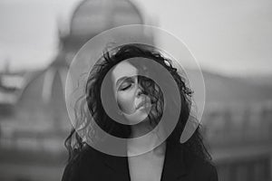 Young attractive brunette in a black jacket stands on the roof with closed eyes against the background of the urban