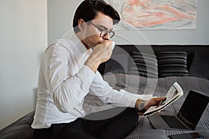 Young attractive brunette man in white shirt and eyeglasses drinking coffee and dreamily reading newspaper on bed with