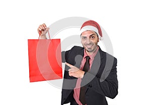 Young attractive businessman in Christmas Santa hat holding and pointing red shopping bag in December and New year sale