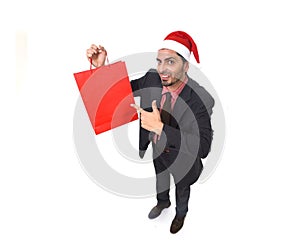 Young attractive businessman in Christmas Santa hat holding and pointing red shopping bag in December and New year sale