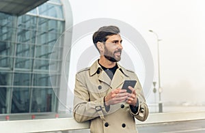 Young Attractive Businessman Standing near Big Modern Office Building. Typing a Message on his Smartphone