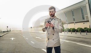Young Attractive Businessman Standing near Big Modern Office Building. Typing a Message on his Smartphone