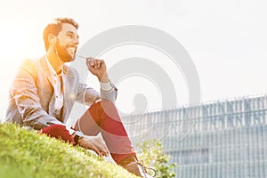 Young attractive businessman talking on smartphone with earphones on while sitting on the grass