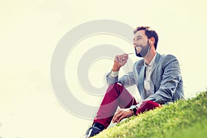 Young attractive businessman talking on smartphone with earphones on while sitting on the grass