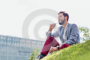 Young attractive businessman talking on smartphone with earphones on while sitting on the grass