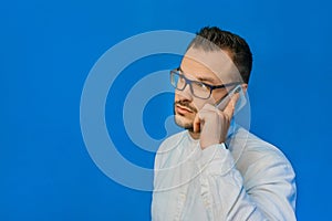 Young attractive businessman in a white shirt and glasses speaks on the phone and smiles on a blue background