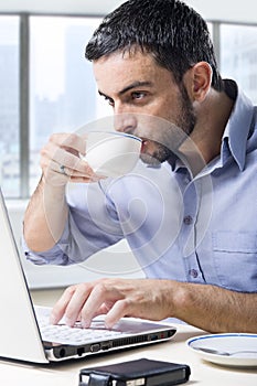 Young attractive businessman working on computer laptop drinking cup of coffee cup sitting at office desk