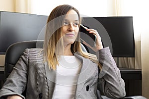 Young attractive businesswoman talking on the phone while sitting in a executive chair with computers on the background.