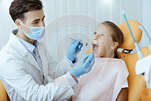 Young attractive caring man in white coat, protective mask and gloves doing medical procedure for dental treatment