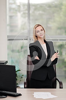 Young attractive Caucasian blond woman in black business suit sits at desk in bright office. Studying paper documents