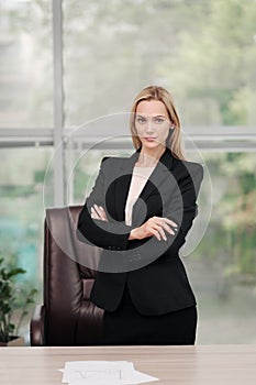 Young attractive Caucasian blond woman in black business suit sits at desk in bright office. Studying paper documents