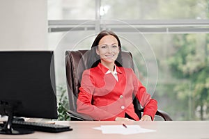 A young attractive Caucasian blond woman in a red business suit sits at a desk in a bright office. Portrait of a