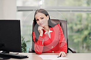 A young attractive Caucasian blond woman in a red business suit sits at a desk in a bright office. Portrait of a