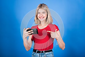 A young and attractive Caucasian blonde girl in casual clothes using a mobile phone and pointing her finger at it.