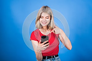 A young and attractive Caucasian blonde girl in casual clothes using a mobile phone and pointing her finger at it.