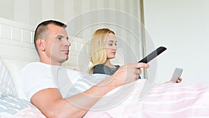 Young attractive caucasian couple in bed. Man watching tv, woman browsing web on digital tablet. Couple in bed not speaking