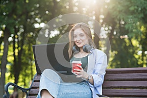 A young and attractive Caucasian girl in headphones sitting on a bench in the park. Using a laptop. Freelance, Network concept.