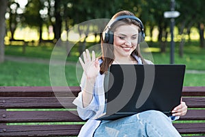 A young and attractive Caucasian girl in headphones sitting on a bench in the park. Using a laptop. Greeting, distance conference.