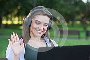A young and attractive Caucasian girl in headphones sitting on a bench in the park. Using a laptop. Greeting, distance conference.