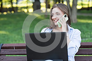 A young and attractive Caucasian girl sitting on a bench in the park. Talking on the phone and using a laptop. Freelance concept.