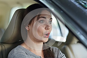 Young attractive caucasian woman behind the wheel driving a car with grimace of astonishment or shock, mouth opened.