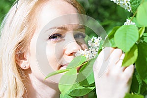 Young attractive caucasian woman in green forest with smile is sniffing blossom of bird-cherry, holding it by hands at sunny day