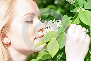 Young attractive caucasian woman in green forest with smile is sniffing blossom of bird-cherry, holding it by hands at sunny day