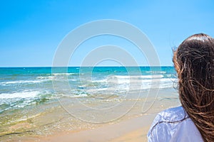 Young Attractive Caucasian Woman with Long Chestnut Hair with Back to Viewer Stands On Sand Beach Looks at Turquoise Sea Horizon