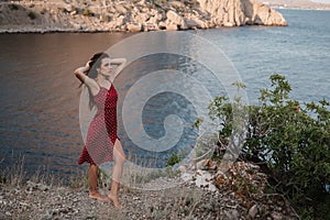 A young attractive Caucasian woman in a red dress at the edge of a cliff near the beach and the sea raised her hands up