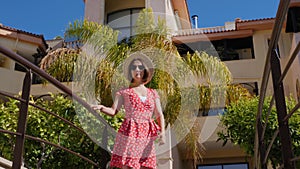 Young attractive caucasian woman wearing red dress, hat and sunglasses looking at the sun with palm trees on the background. Slow