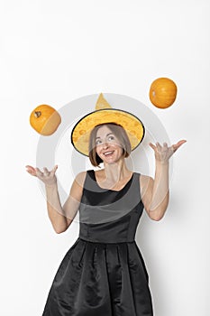 Young attractive and cheerful woman in black beautiful dress and witch hat throwing up two pumpkins isolated on white background