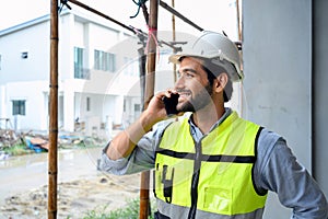 Young attractive construction smiling bearded using smartphone calling to client, worker in vest with white helmet standing on