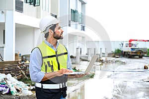 Young attractive construction smiling bearded in vest with white helmet working with laptop, standing on building construction