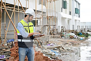 Young attractive construction smiling bearded in vest with white helmet working with laptop, standing on building construction