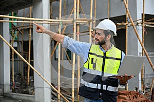 Young attractive construction smiling bearded in vest with white helmet working with laptop, standing on building construction