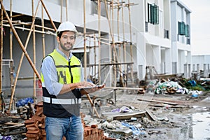 Young attractive construction smiling bearded in vest with white helmet working with laptop, standing on building construction