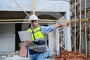 Young attractive construction smiling bearded in vest with white helmet working with laptop, standing on building construction