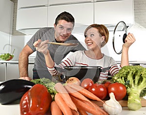 Young attractive couple at home kitchen with man tasting vegetable stew cooked by her wife smiling happy