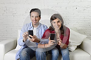 Young attractive couple together at home couch with arms interlaced using mobile phone compulsively