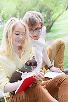 Young attractive couple wearing glasses is working or studying with laptop book note and pen sitting on blanket in green park at s