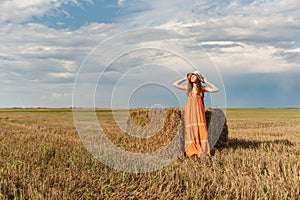 A young attractive curly rural woman in a retro vintage dress and a hat stretching stands near a stack of harvested wheat straw in