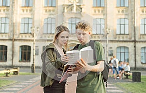 Young attractive cute students couple standing in front of the university reading an interesting book. Two concentrated amazed