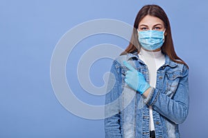 Young attractive dark haired fashionable girl wearing medical mask and latex gloves points aside with her index finger, looking at